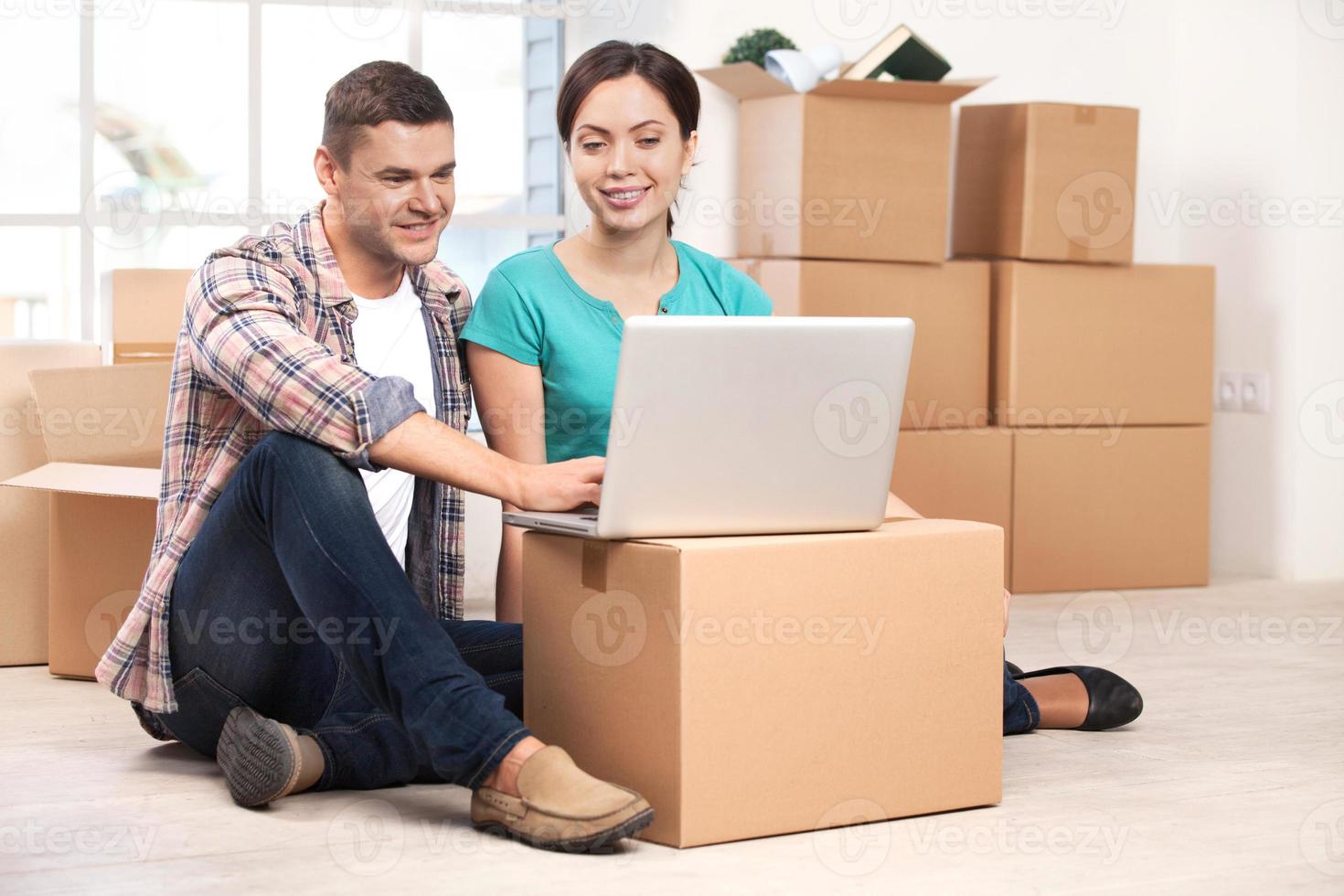 Surfing the net together. Cheerful young couple sitting on the floor and working on laptop while cardboard boxes laying on the background photo