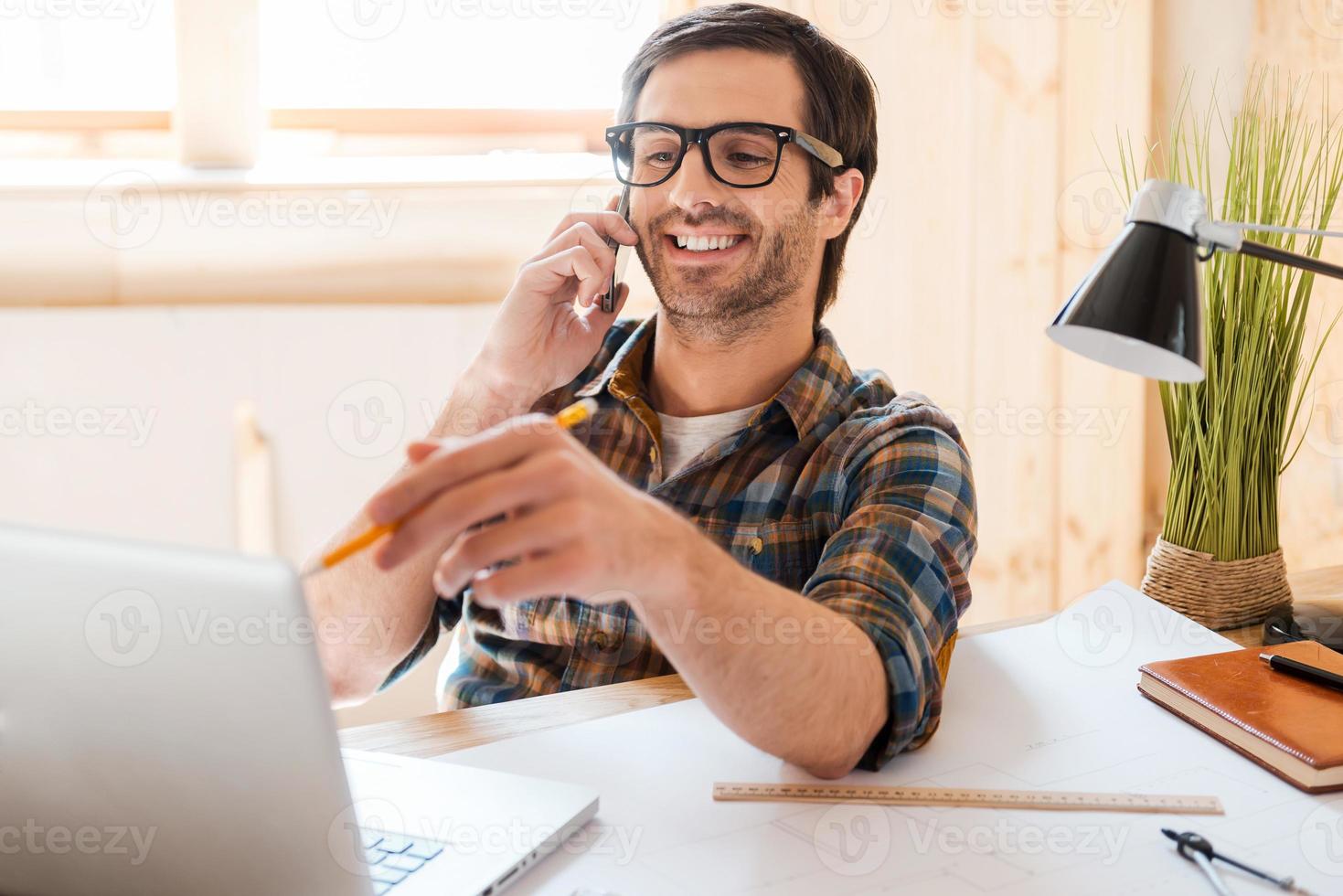 Creative worker. Cheerful young man talking on mobile phone and pointing at the laptop while sitting at his working place photo