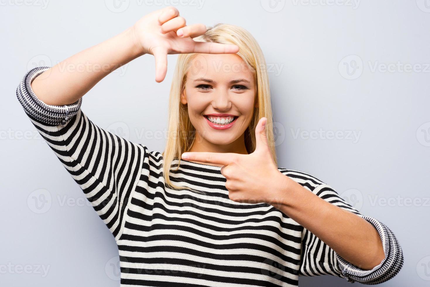 marco de dedo gesticular. hermosa joven rubia mirando a la cámara y haciendo gestos con el marco del dedo mientras se enfrenta a un fondo gris foto