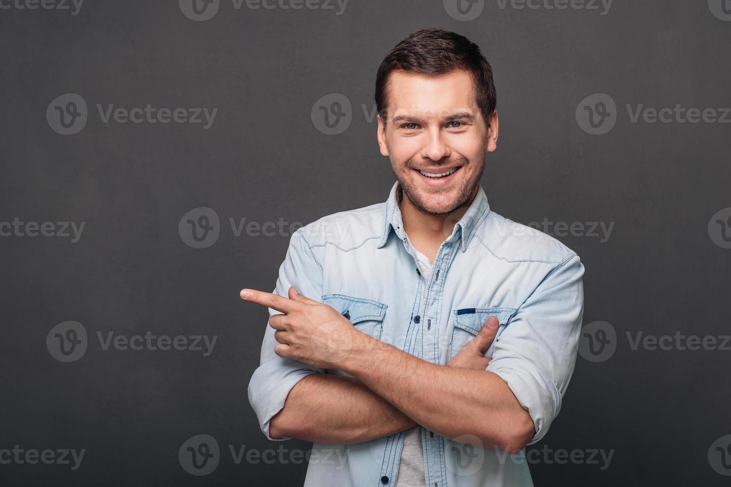 Your answer is here Cheerful young man pointing at copy space and looking at camera with smile while standing against grey background photo