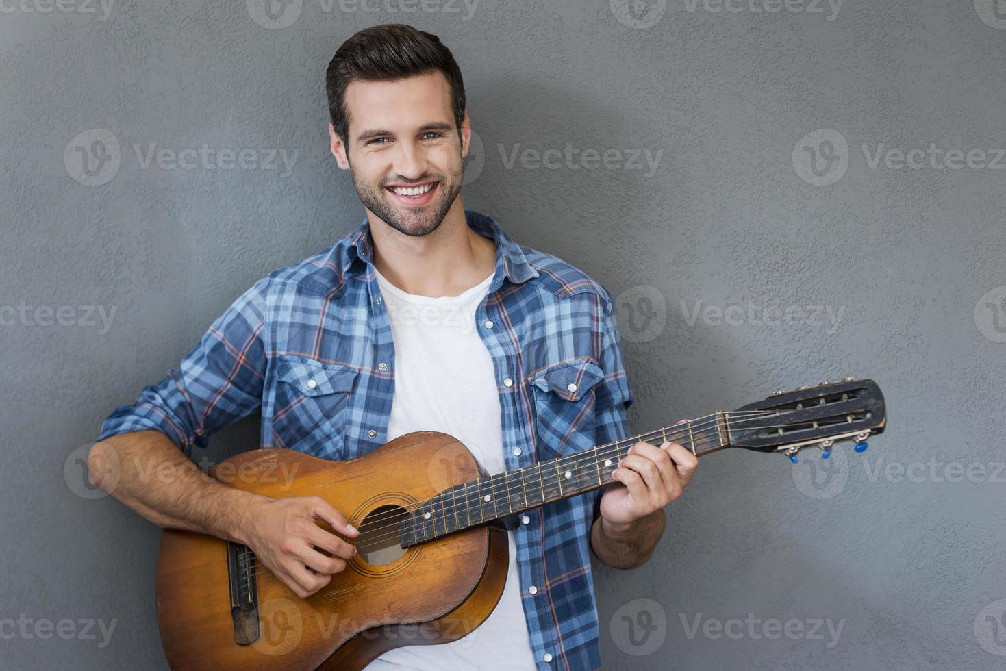 Young man with guitar. Happy young man playing the guitar and looking at camera while standing against grey background photo