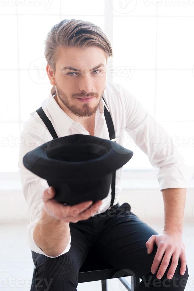 In style. Handsome young man in shirt and suspenders stretching out hand with hat in it and looking at camera while sitting on the chair photo