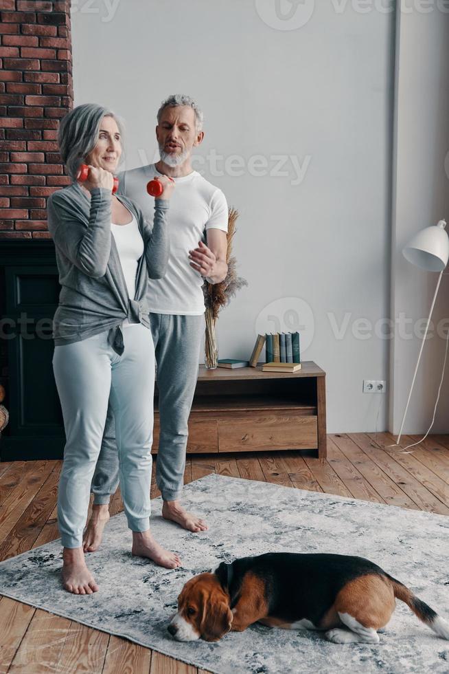 Full length of senior couple in sports clothing exercising using hand weight and smiling while spending time at home photo