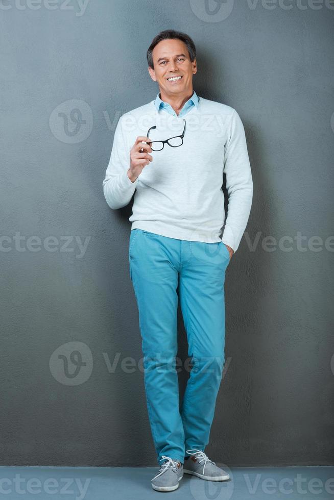 Smart and casual. Full length of cheerful mature man holding hand in pockets and smiling at camera while standing against grey background photo