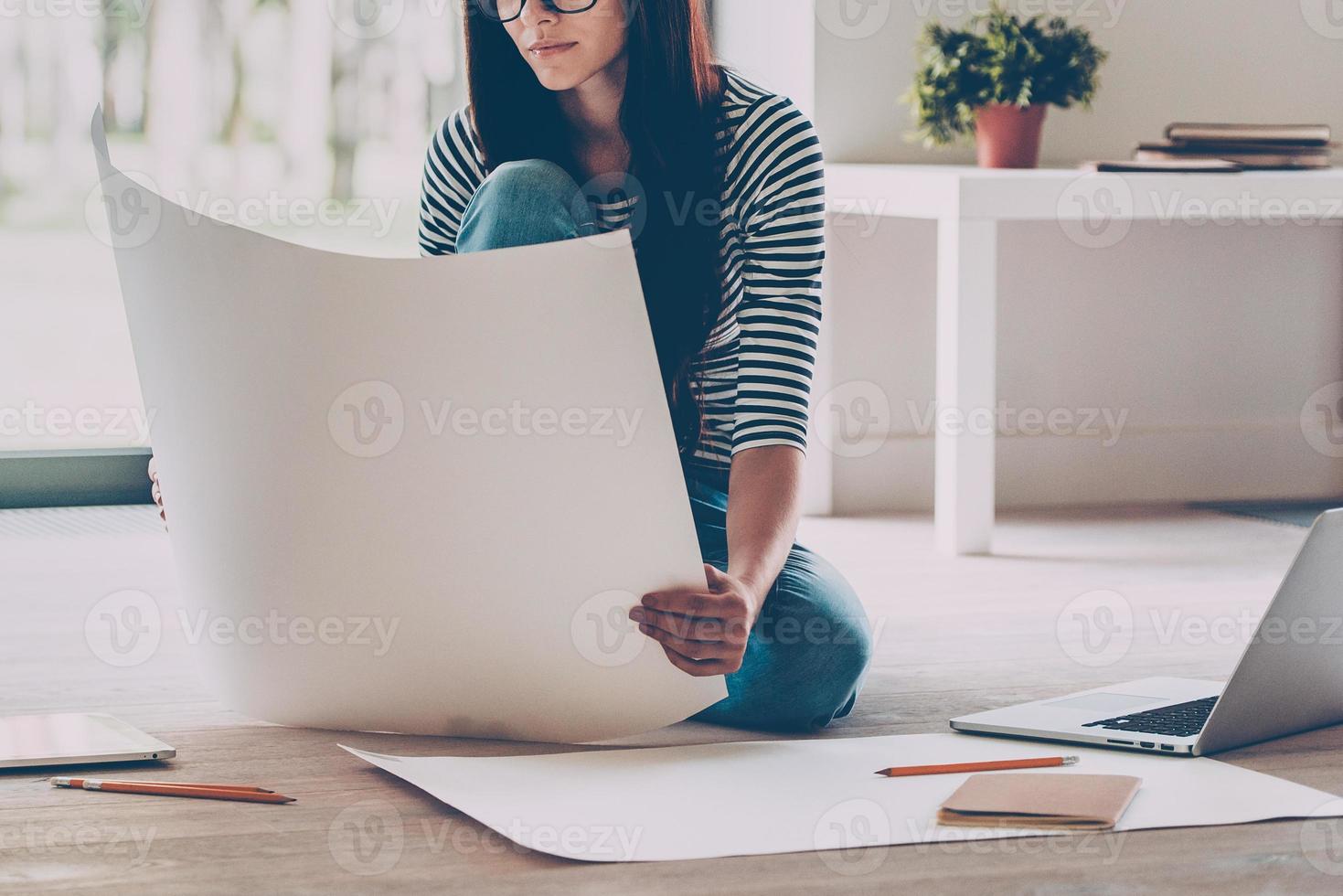 Confident architect. Close-up of confident young beautiful woman examining blueprint while sitting on the floor at home photo