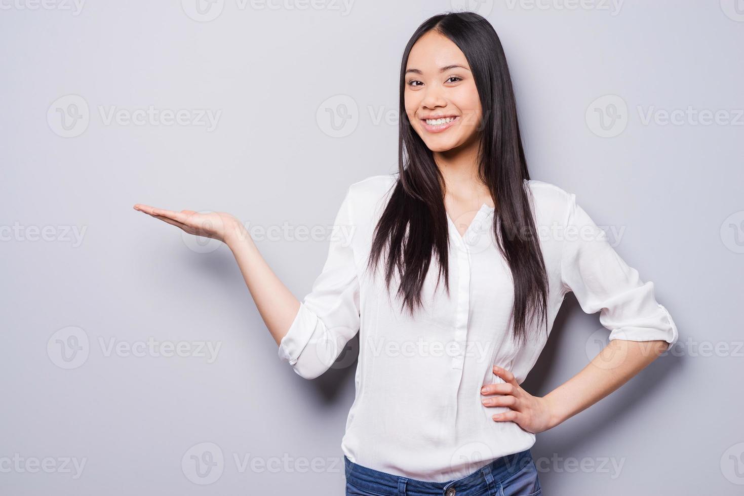 copie el espacio en su mano. alegre joven mujer asiática sosteniendo un espacio de copia y sonriendo mientras está de pie contra el fondo gris foto