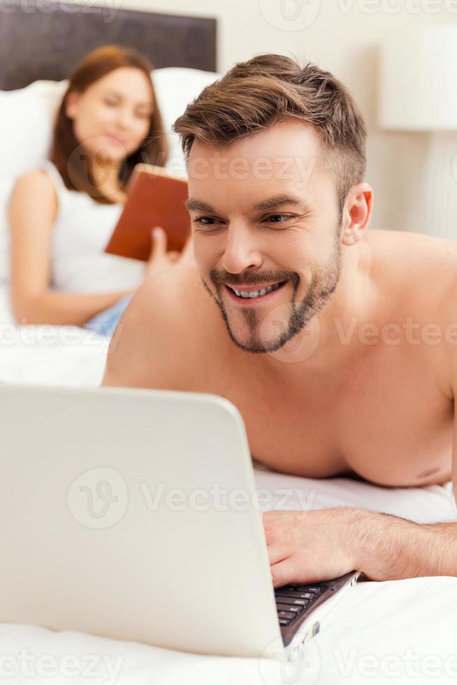 Spending leisure time in bed. Handsome young and shirtless man lying in bed and working on laptop while woman reading a book in the background photo