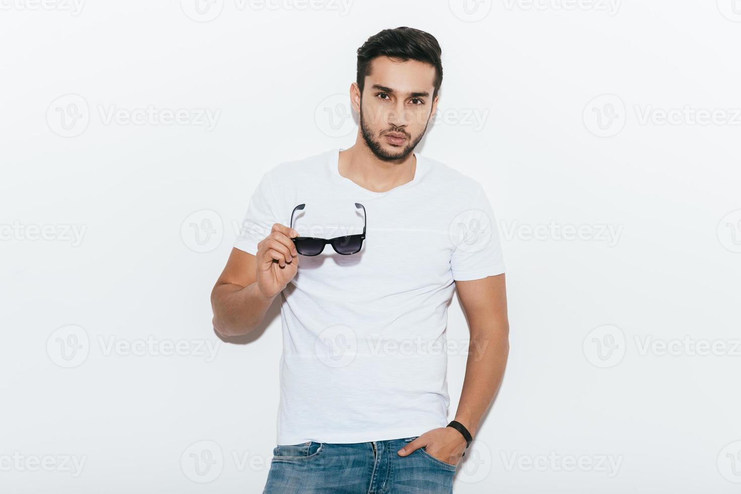 encantador guapo. apuesto joven indio con gafas de sol y mirando a la cámara mientras está de pie contra el fondo blanco foto