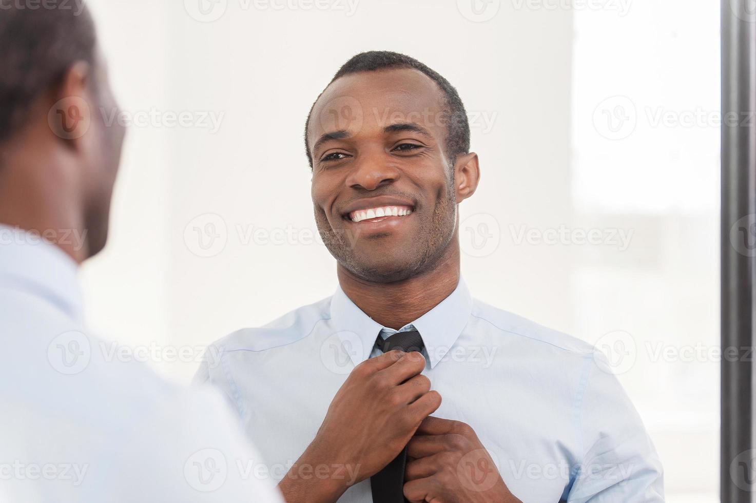 Confident about his look. Young African man adjusting his necktie while standing against mirror photo