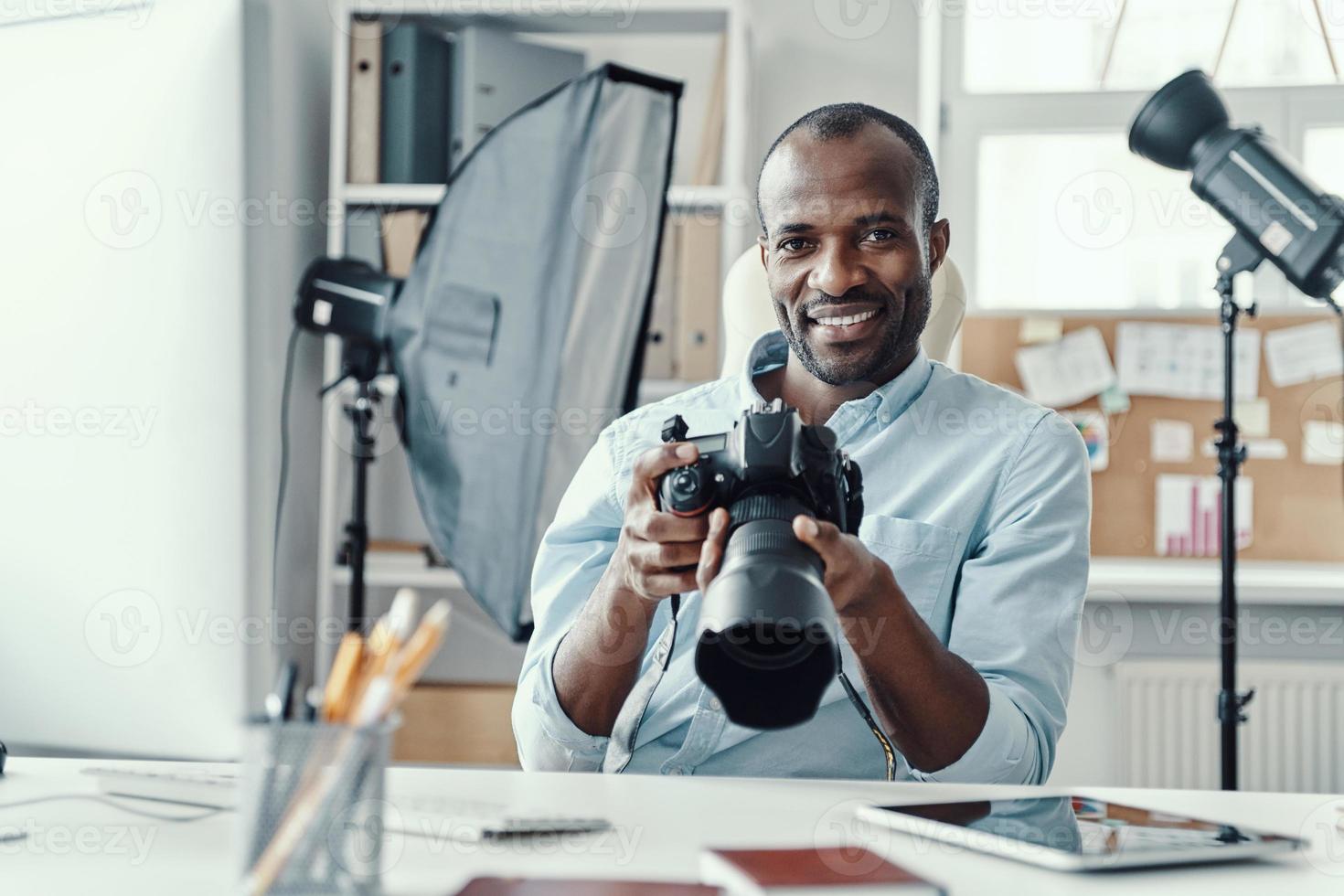 apuesto joven africano sosteniendo una cámara digital y sonriendo mientras trabajaba en la oficina moderna foto