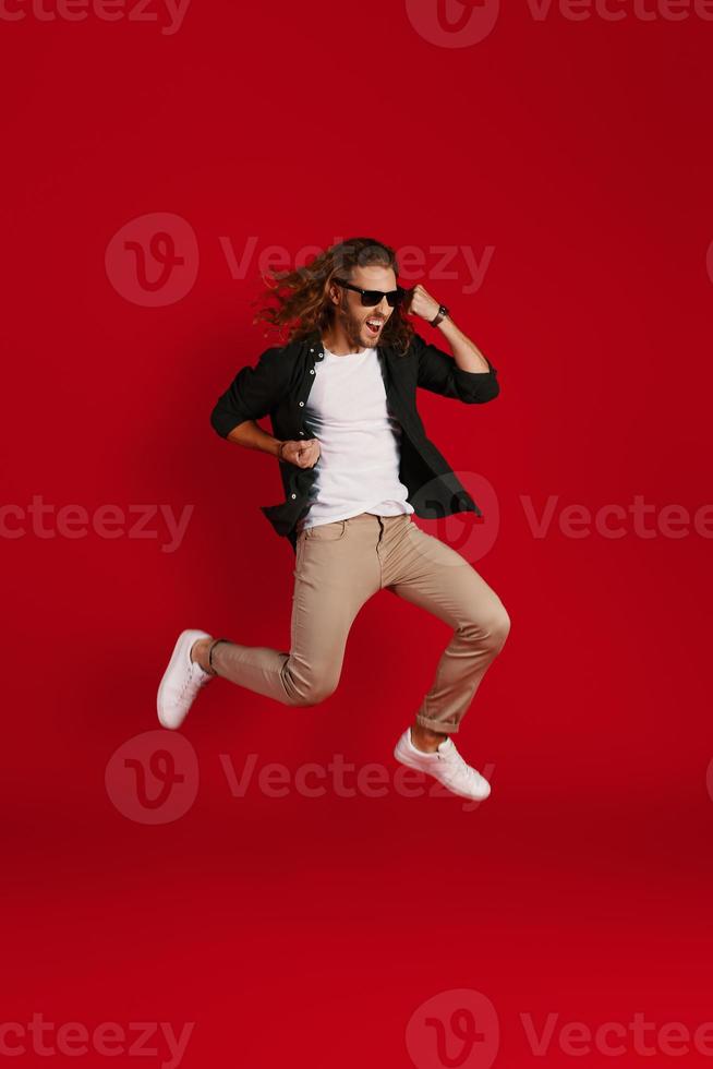 Full length of happy young man in casual clothing smiling and gesturing while hovering against red background photo