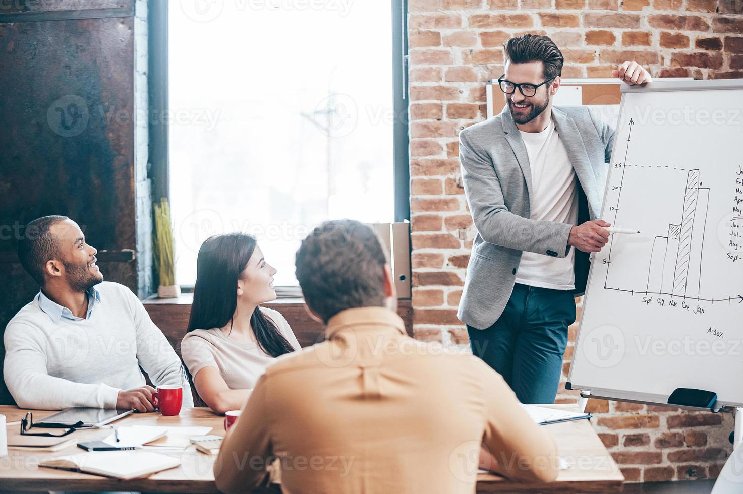 We have some great news Handsome young man in glasses standing near whiteboard and pointing on the chart while his coworkers listening and sitting at the table photo