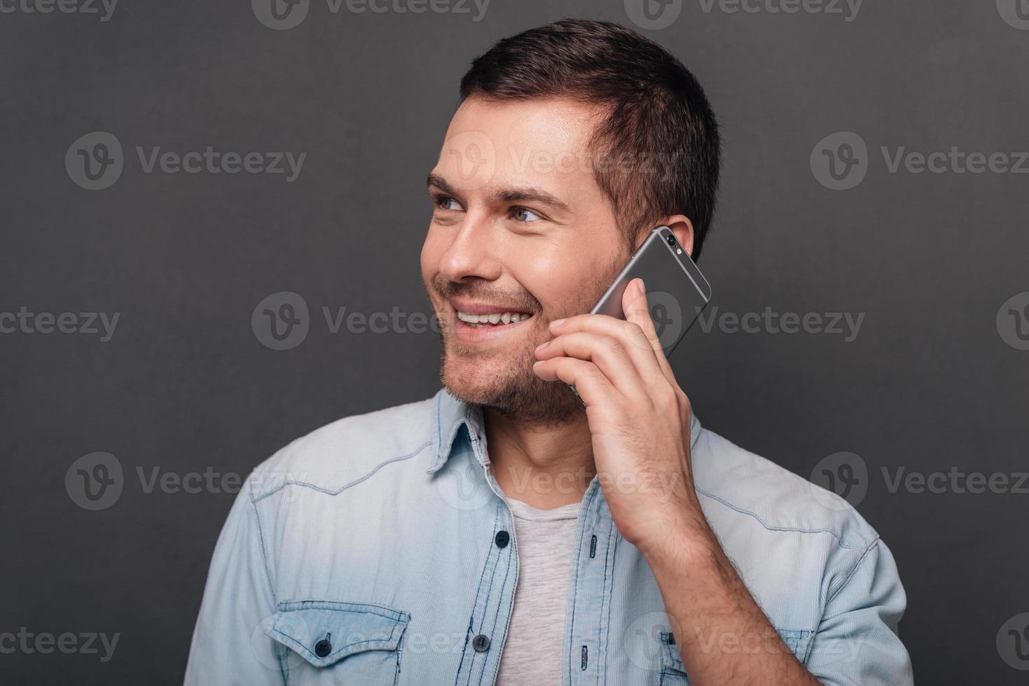 Nice to hear you Cheerful young man talking on mobile phone and looking away with smile while standing against grey background photo