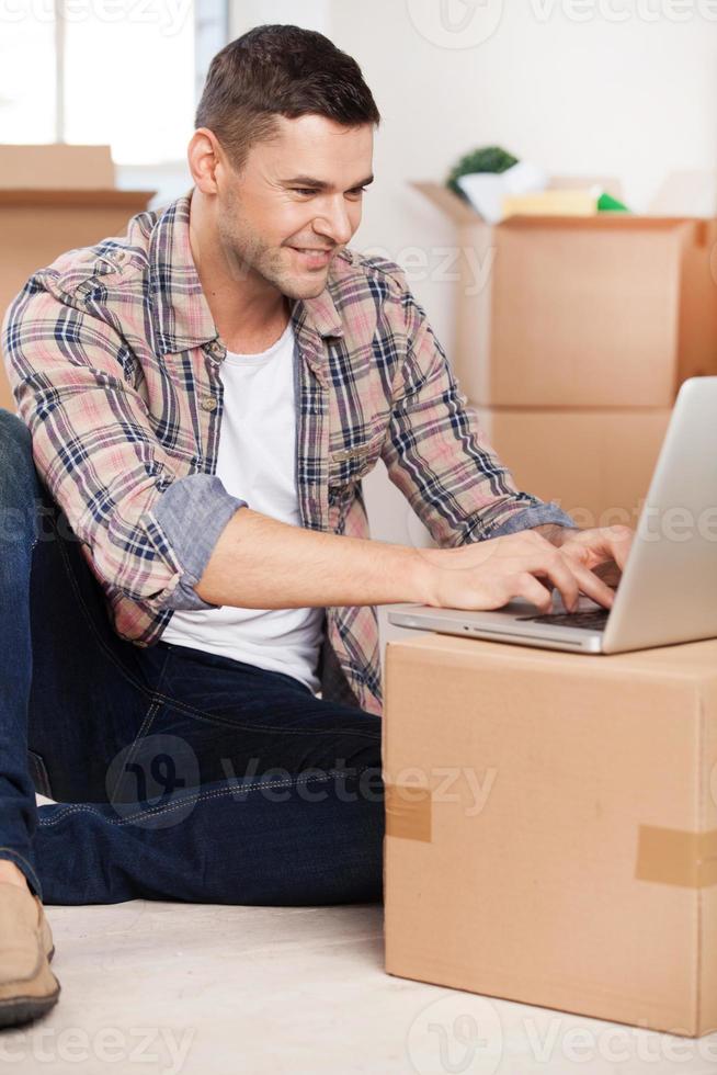 Surfing web in a new house. Handsome young man sitting on the floor and typing something on laptop while cardboard boxes laying on the background photo