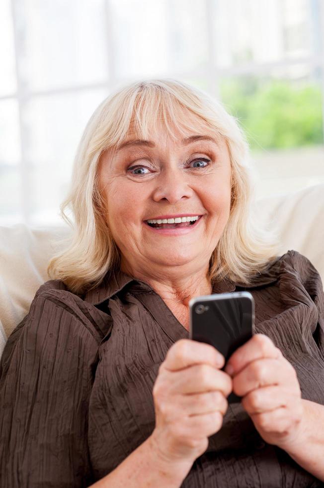 Senior woman with mobile phone. Cheerful senior woman holding mobile phone and smiling while sitting in a chair photo