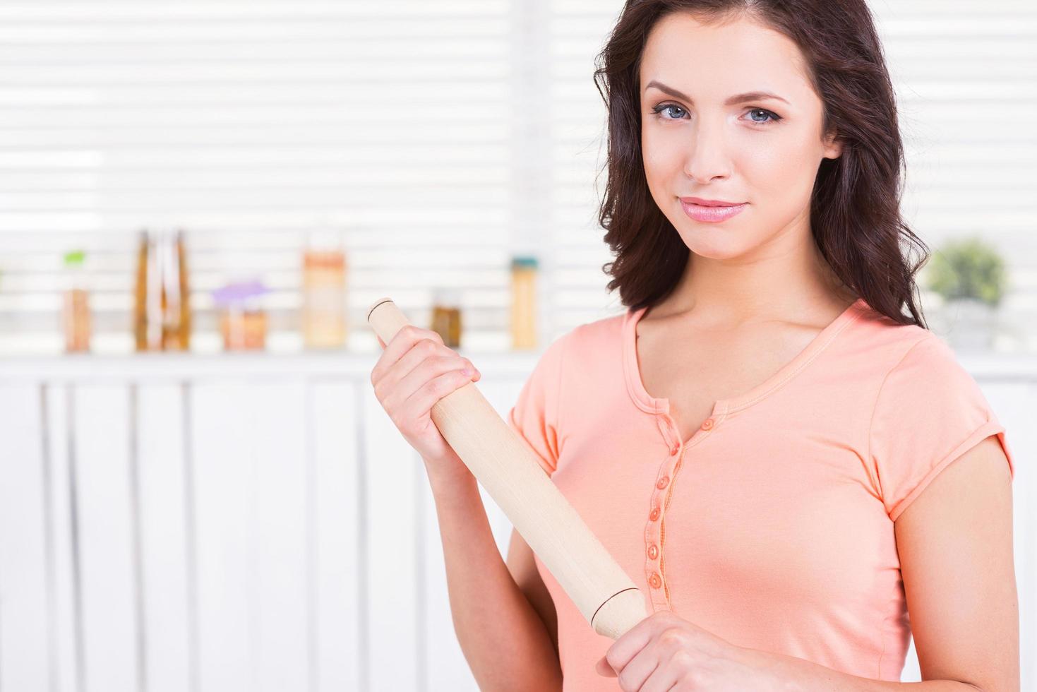 Housewife with rolling pin. Beautiful young woman holding rolling pin and looking at camera while standing in a kitchen photo