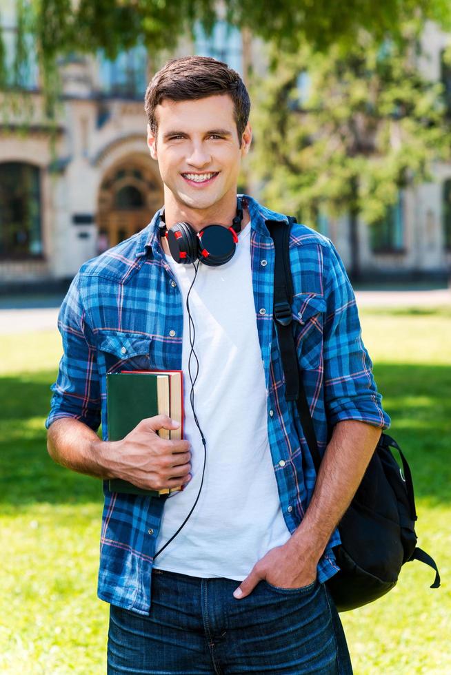 Ready to study. Handsome young man holding books and smiling while standing in front of his university photo