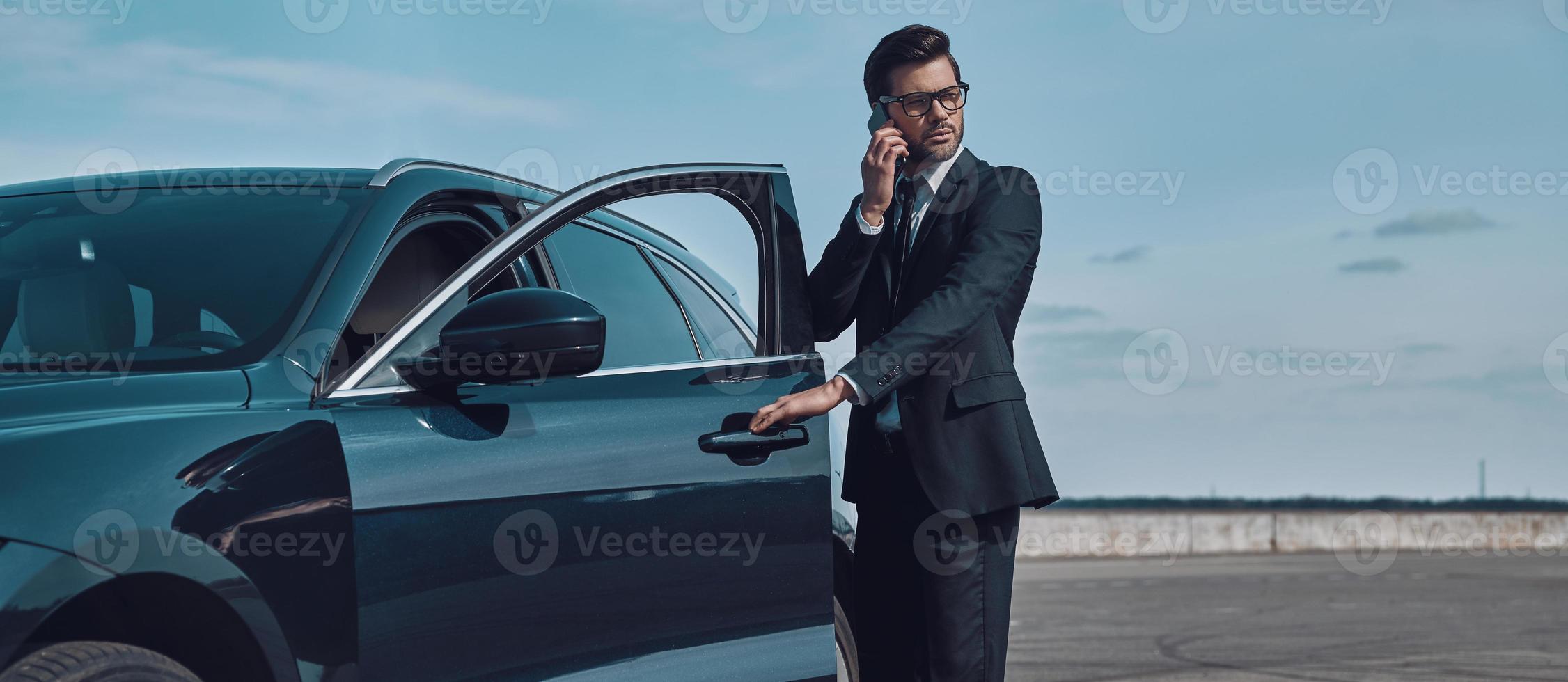 experto en negocios confiado. toda la longitud de un apuesto joven hombre de negocios hablando por teléfono mientras entra en su auto al aire libre foto