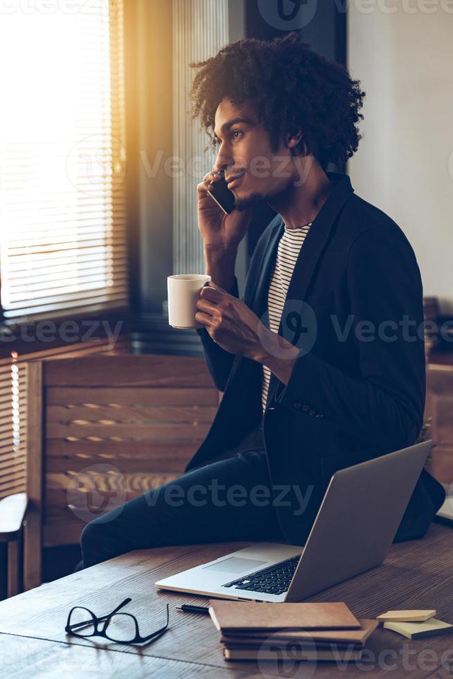 I have some great news for you Side view of young handsome African man talking on mobile phone and holding coffee cup while sitting on table at his working place photo