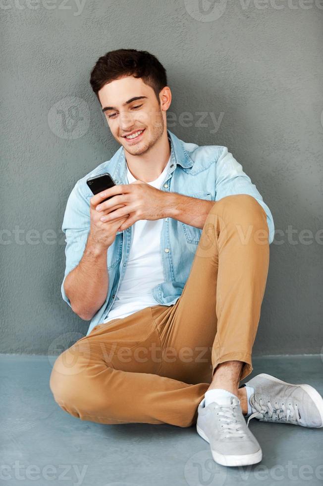 Sending message to friend. Joyful young man holding mobile phone and smiling while sitting against grey background photo