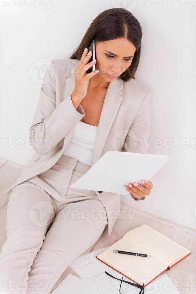 Busy working. Top view of confident young businesswoman examining document and talking on the mobile phone while sitting on the floor photo