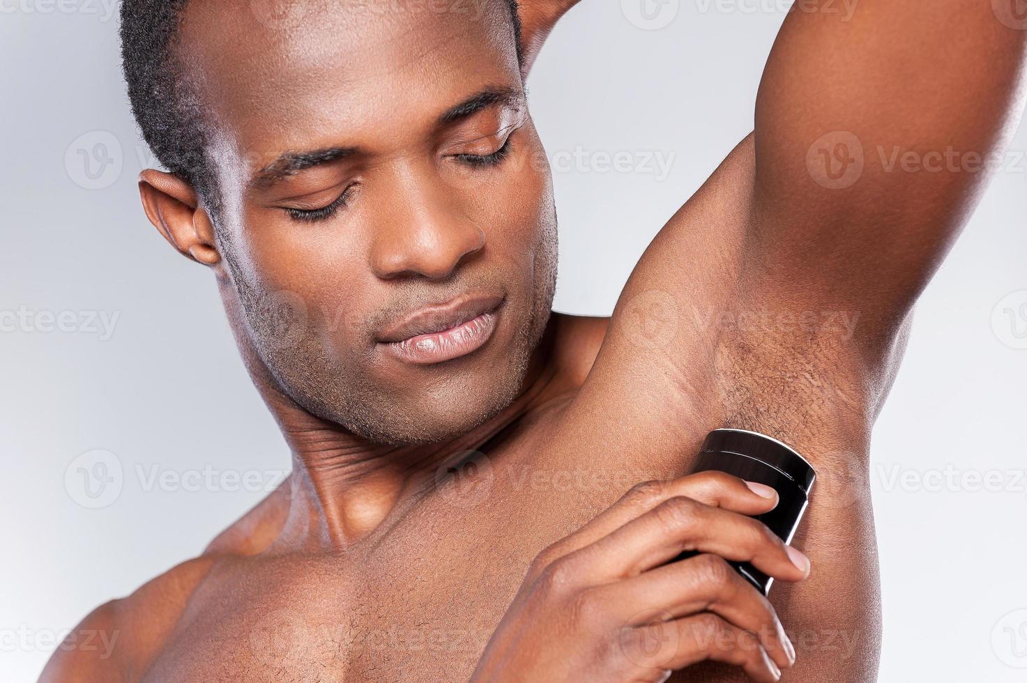 Always clean and fresh. Young shirtless African man using dry deodorant while standing against grey background photo