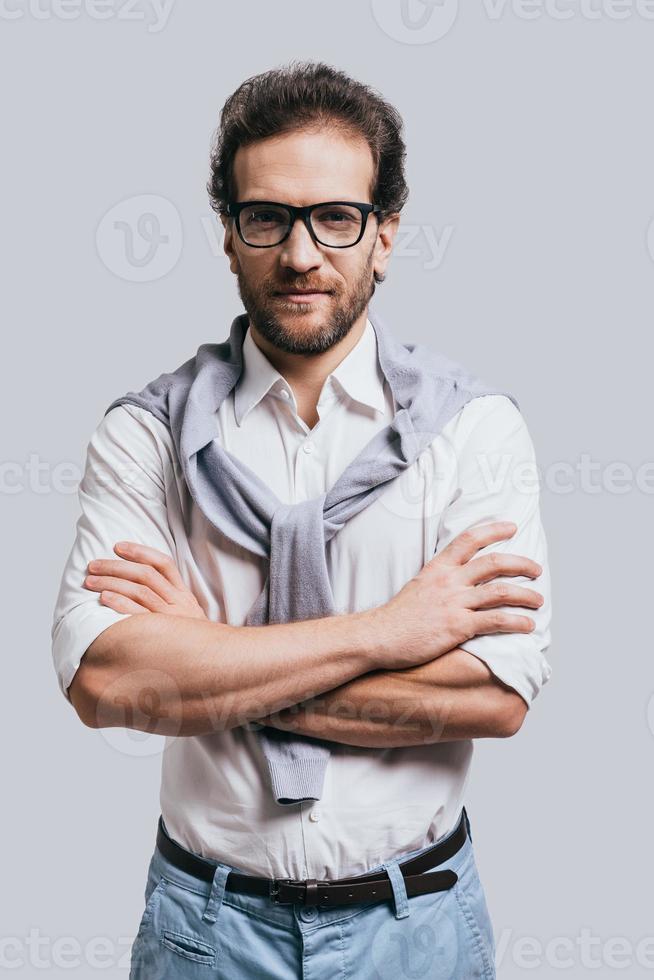 Confidence and style. Handsome young man keeping arms crossed and looking at camera while standing against grey background photo