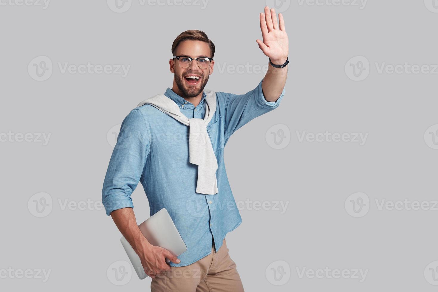 Good looking young man in eyeglasses holding his digital tablet and gesturing while standing against grey background photo