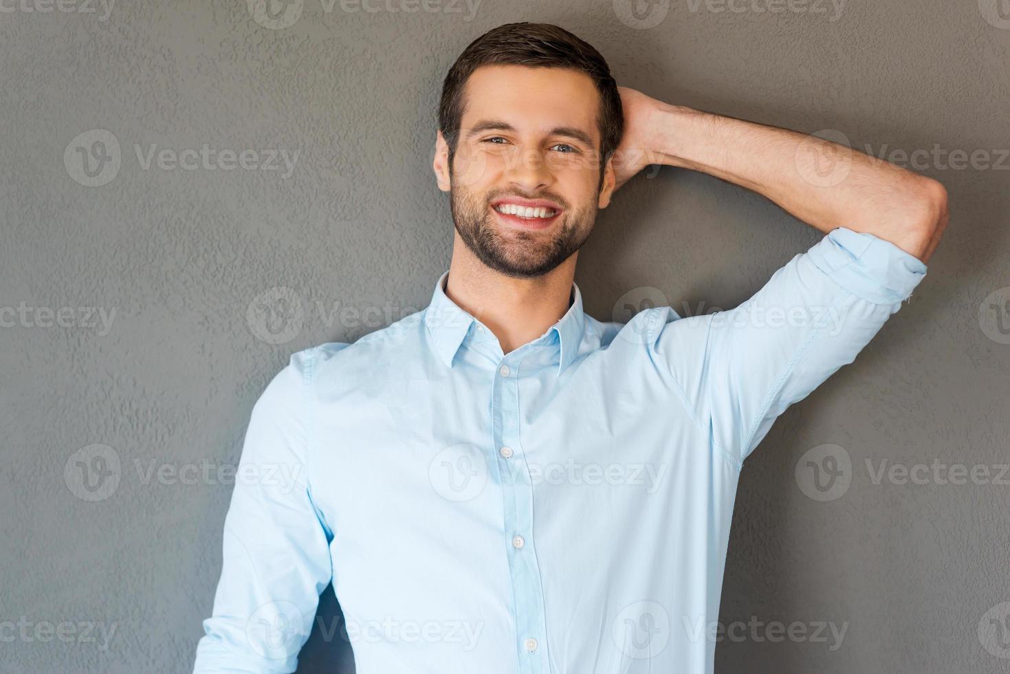 Feeling satisfied and relaxed. Handsome young man in shirt touching his head and smiling at camera while standing against grey background photo