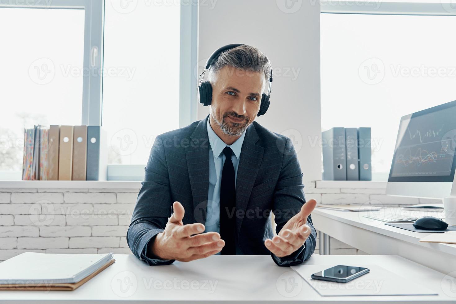 Handsome businessman in headphones smiling and gesturing while sitting at his working place photo