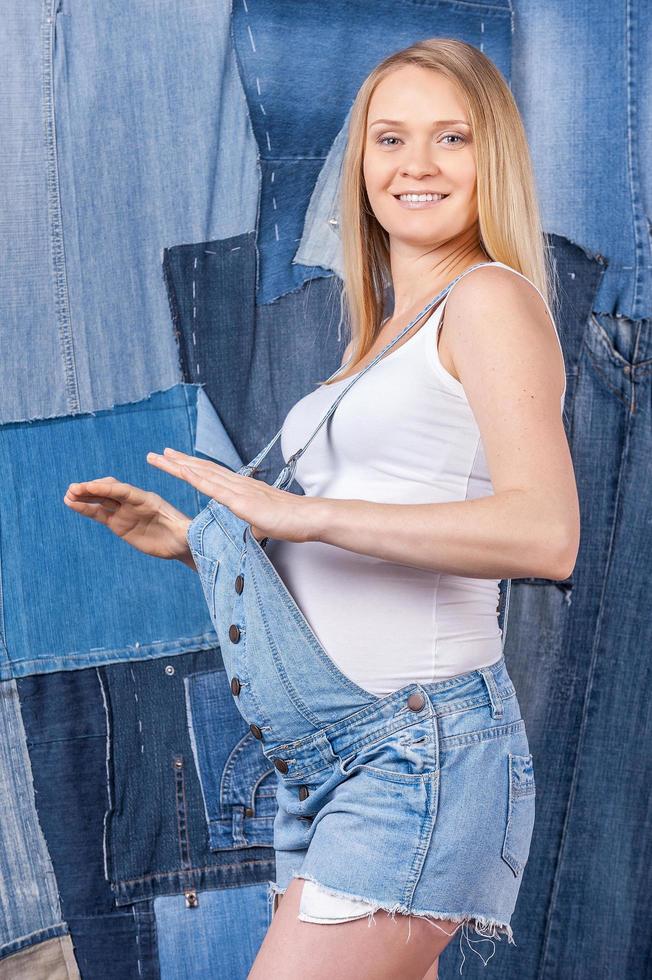 siempre luciendo a la moda. vista lateral de una hermosa mujer embarazada con ropa de jeans posando mientras está de pie contra el fondo de los jeans foto