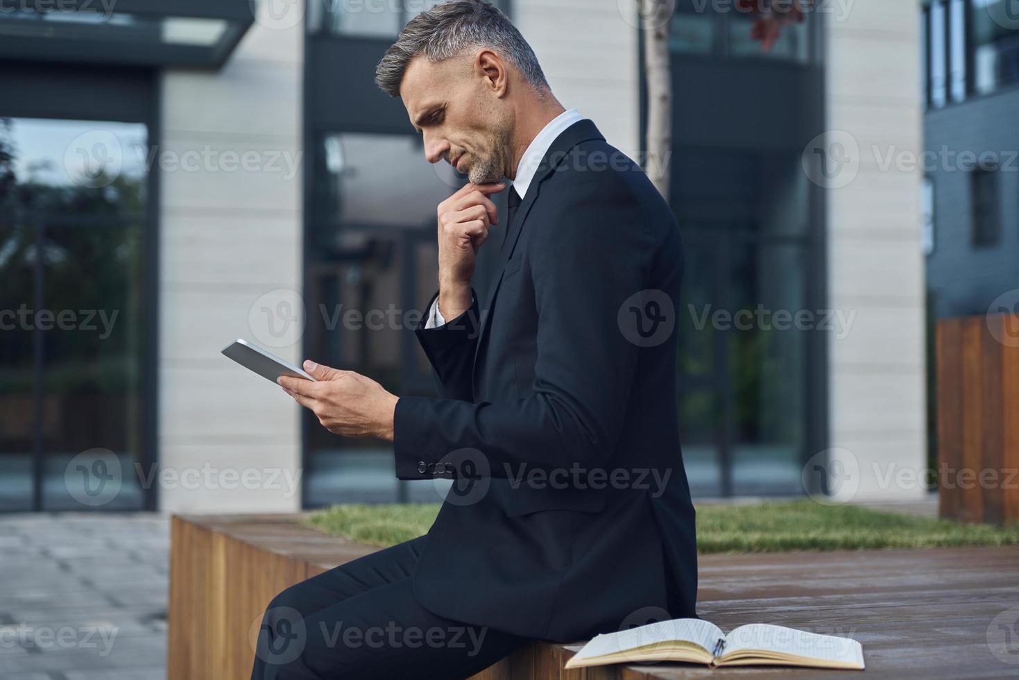 Confident mature businessman using digital tablet while sitting outdoors near office building photo