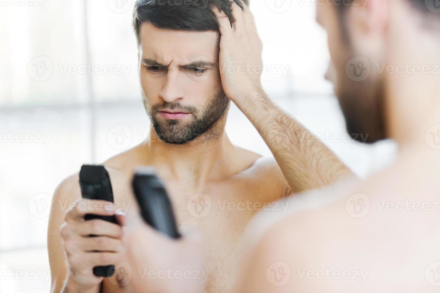 Bad shaver. Rear view of frustrated young man holding an electric shaver and looking at it while standing in front of the mirror photo