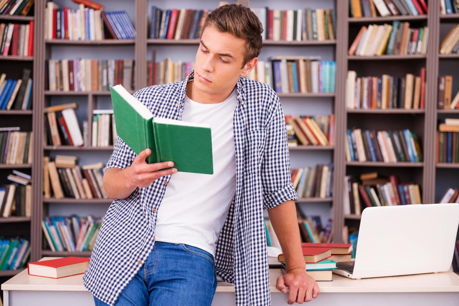 Reading his favorite book. Confident young man reading book while leaning at the desk in library photo