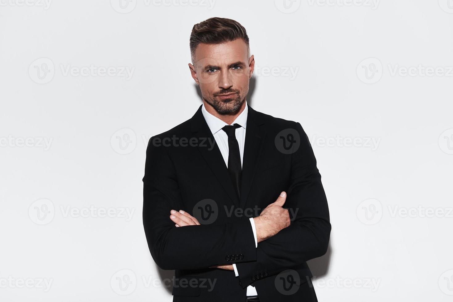 Confident business expert. Handsome young man in full suit keeping arms crossed and looking at camera while standing against white background photo