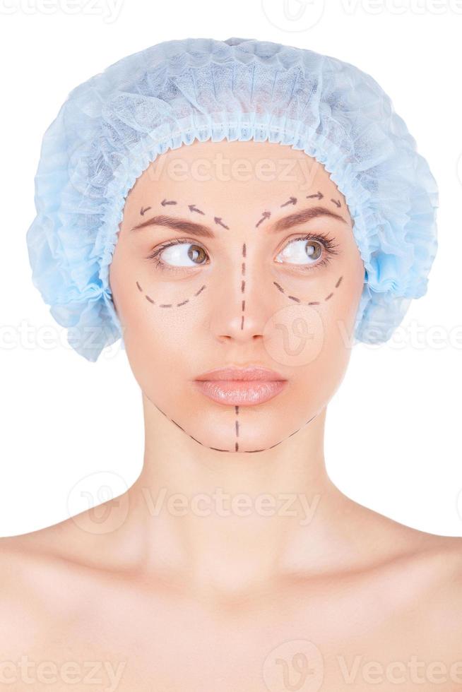 Ready for plastic surgery. Beautiful young woman in medical headwear and sketches on face looking away while isolated on white photo