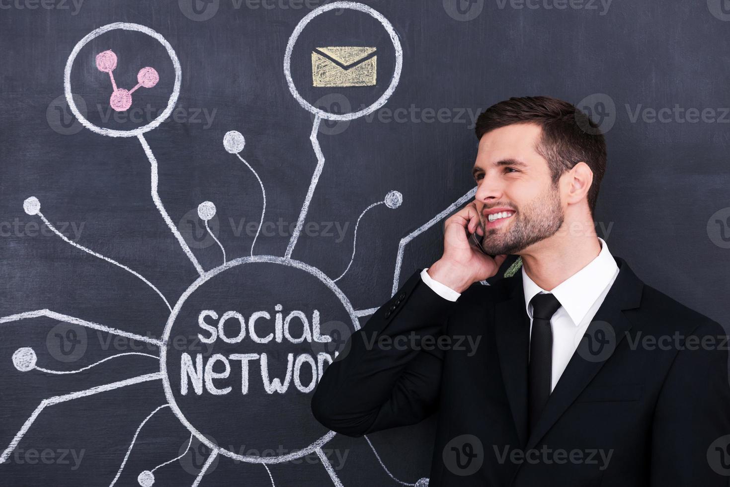 Active social life. Handsome young man speaking on telephone while standing against social network chalk drawing on blackboard photo