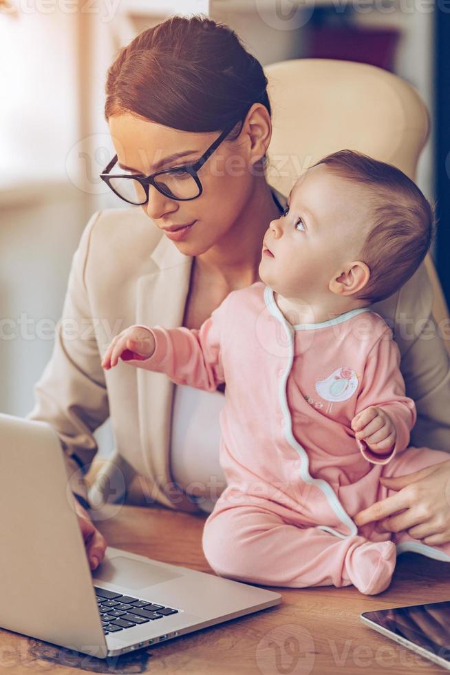 madre trabajadora a tiempo completo. niñita mirando hacia arriba mientras se sienta en el escritorio de la oficina con su madre en la oficina foto