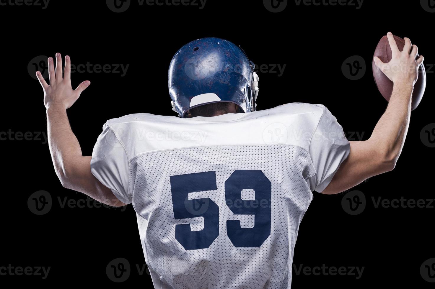 apoyo de la afición. vista trasera del jugador de fútbol americano sosteniendo una pelota de fútbol manteniendo los brazos levantados mientras está de pie contra el fondo negro foto