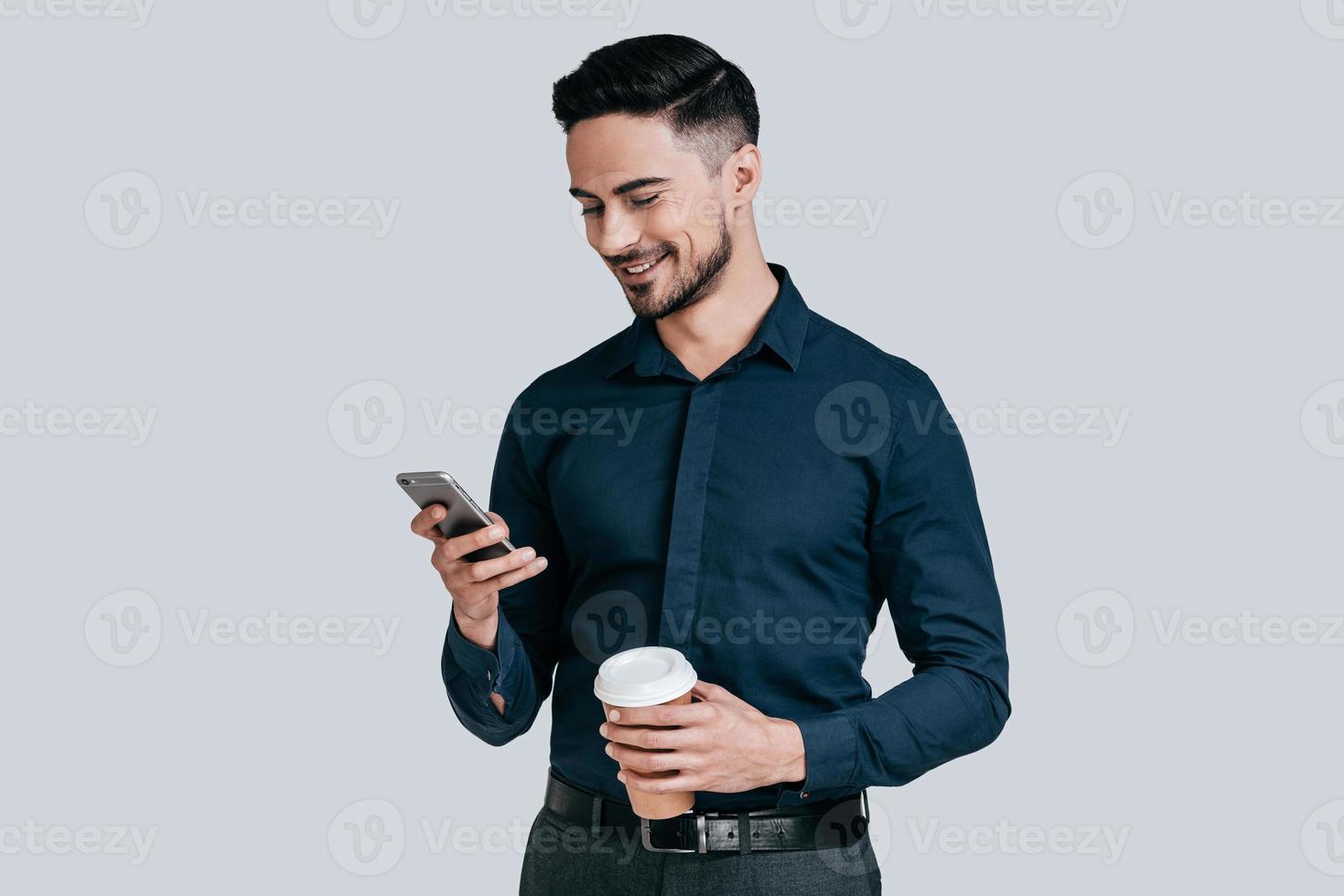 Nice chat. Good looking young man holding smart phone and looking at it with smile while standing against grey background photo