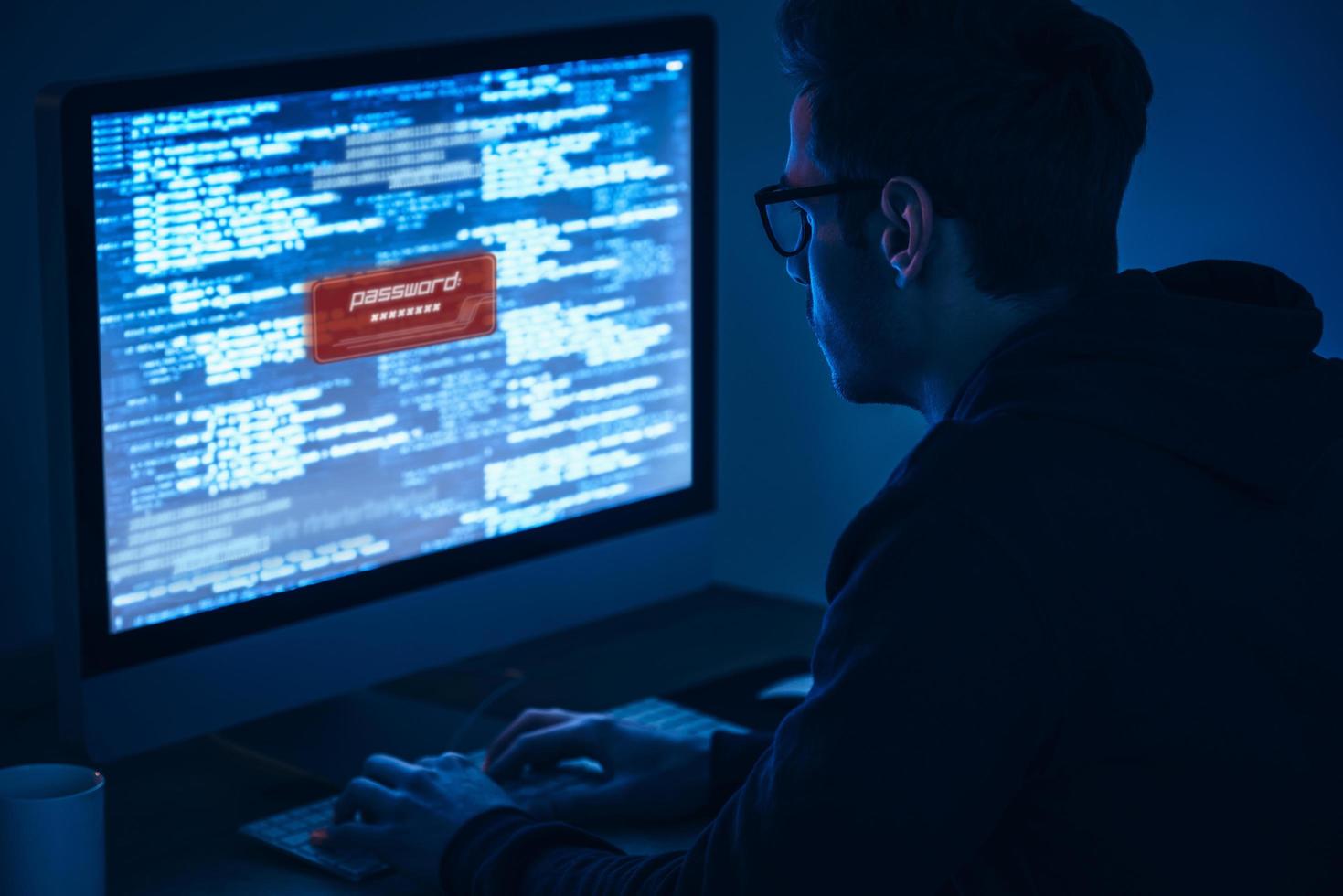 Computer security. Rear view of young man typing and looking at computer monitor while sitting at the table in dark room photo