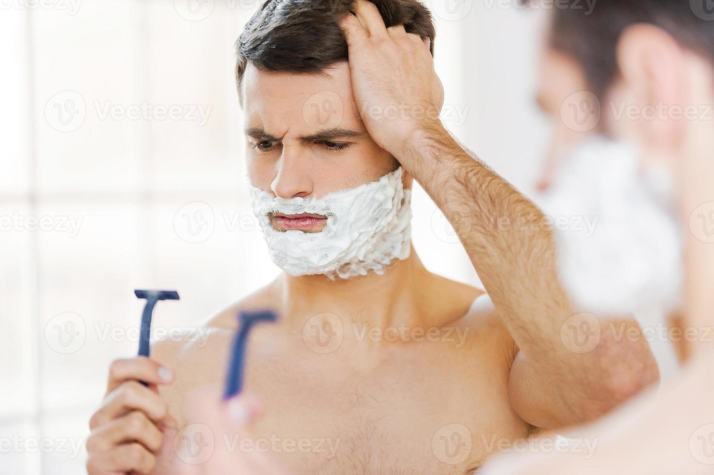 Bad shaver. Rear view of frustrated young shirtless man with shaving cream on his face holding a razor and looking at it while standing in front of the mirror photo