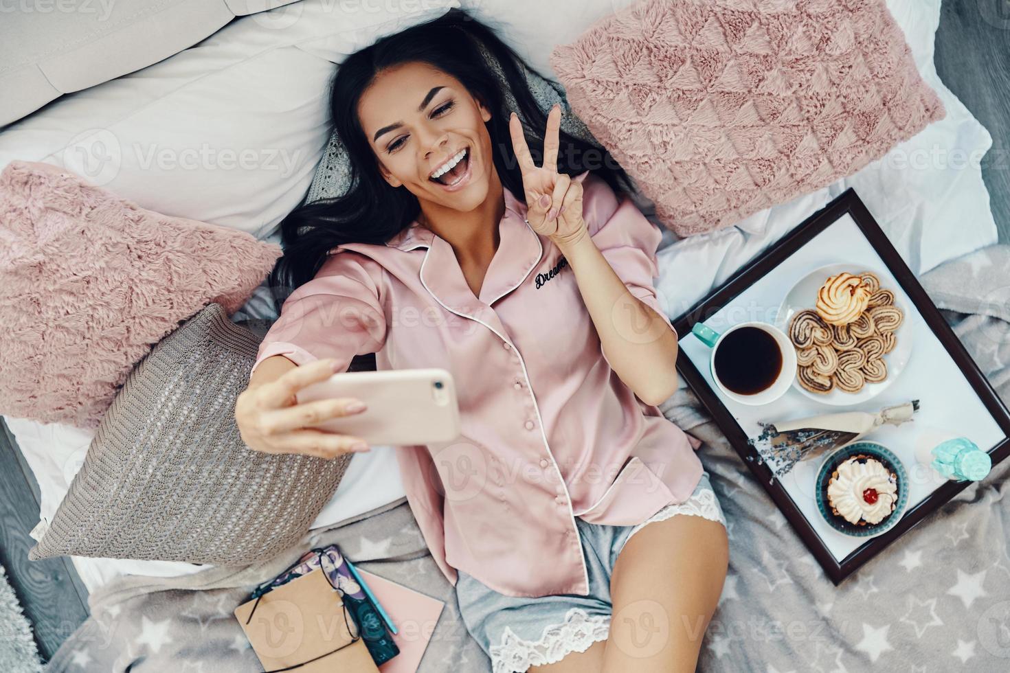 Top view of beautiful young woman in pajamas taking selfie and smiling while resting in bed at home photo