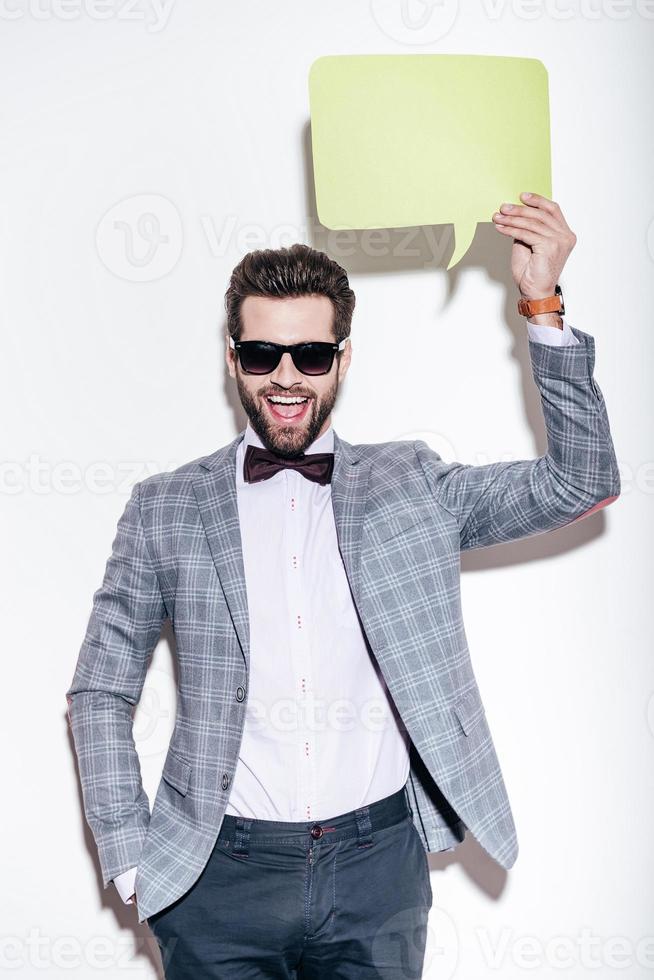 That is what I am saying. Handsome young cheerful man in suit looking at camera with smile and holding green speech bubble upon his head while standing against white background photo