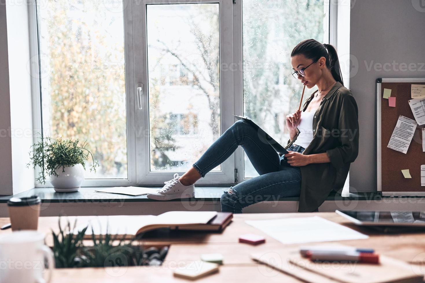 Full concentration at work. Concentrated young woman working while sitting on the window sill in the office photo