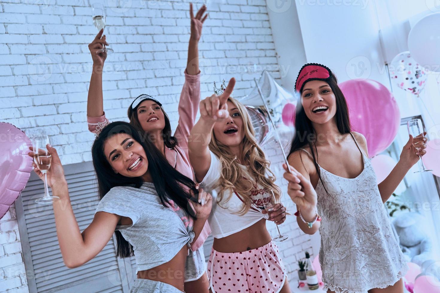 Real fun Four attractive young smiling women in pajamas drinking champagne while having a slumber party in the bedroom photo