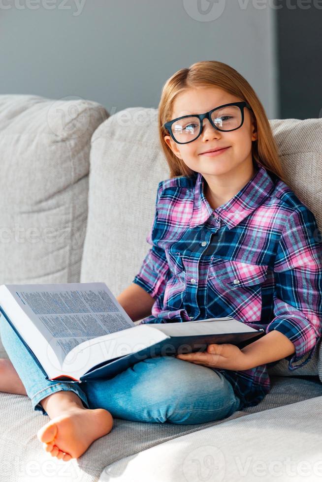 Little cutie with book. Cheerful little girl in glasses holding book and looking at camera with smile while sitting on the couch in lotus position at home photo