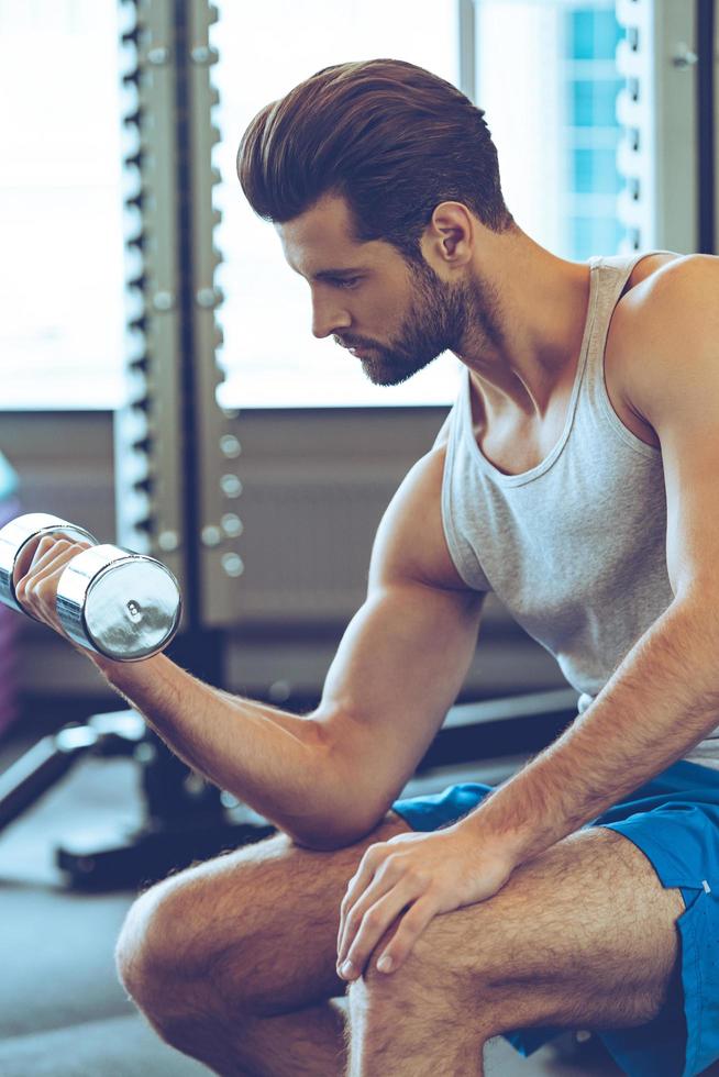 Nice burn in bicep. Side view of confident young handsome man in sportswear exercising with dumbbell at gym photo