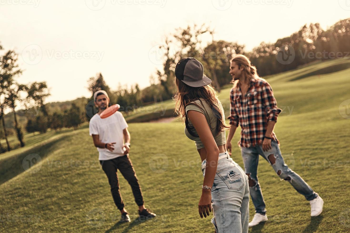Group of young people in casual wear throwing plastic disk while spending carefree time outdoors photo