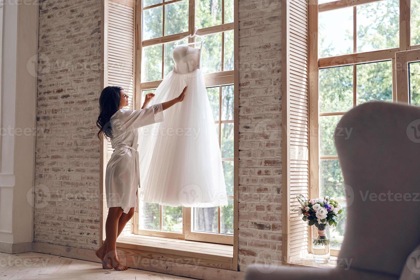 se ve bien. toda la longitud de una hermosa joven con albornoz de seda tocando su vestido de novia mientras está de pie cerca de la ventana foto